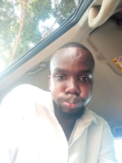 A dark-skinned adult male is depicted in a close-up, chest-up shot, situated within a vehicle. He has a close-cropped haircut and short facial hair around his chin and mouth. He wears a light-colored, collared shirt. His eyes are directed forward, with a neutral expression and slightly pursed lips. Above and behind him, an open sunroof reveals green tree foliage and bright sunlight, indicating an outdoor, daytime setting. Visible interior components of the vehicle include a cream-colored ceiling panel, a light-colored grab handle, and a partially visible yellow warning label with black text (likely an airbag safety sticker). Bright light illuminates the subject from above. The image contains no discernible landmarks or signs to independently verify its location as Makadara/Mji Wa Kale ward, Kenya.