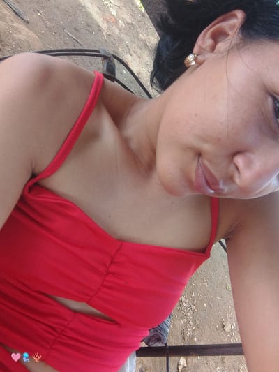 A woman, with dark hair and light skin, is seen in a close-up, low-angle shot. She is wearing a bright red, sleeveless top with thin straps. Her right ear is adorned with a gold hoop earring. Her mouth is slightly open, with her tongue visible. The background consists of dry, dusty ground with scattered pebbles and some dry vegetation. Part of a metal railing is visible in the lower portion of the image. The image appears to have been taken outdoors, possibly in a rural or semi-urban setting.