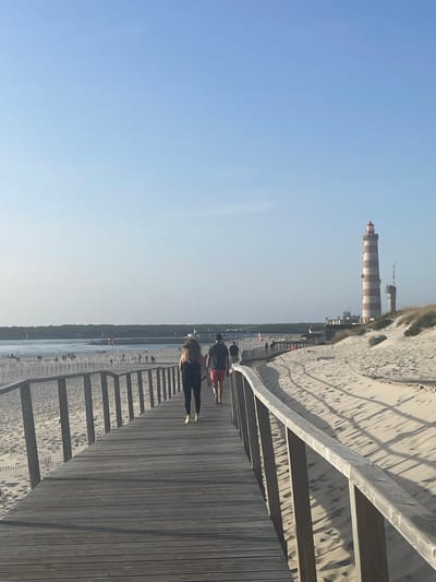 The image shows a wooden boardwalk leading to a sandy beach in Gafanha da Nazaré, Portugal. A lighthouse with red and white stripes stands tall in the distance, accompanied by a smaller tower. The sky is clear and blue. Several people are walking on the boardwalk and the beach. Two individuals are in the foreground, walking away from the camera on the boardwalk. One is wearing black leggings and a black tank top, while the other is wearing red shorts and a black t-shirt. The beach is dotted with sunbathers and beachgoers. The environment appears to be a coastal area with dunes and vegetation. The scene captures a typical day at the beach with people enjoying the scenery and activities.
