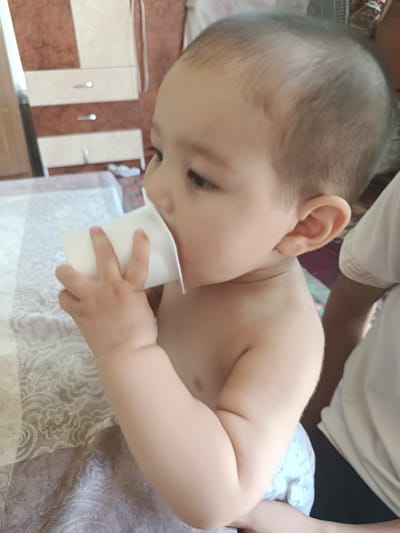 A toddler is sitting on a bed, holding a white plastic cup to their mouth and drinking. The toddler is partially visible from the chest up, wearing a diaper. The background shows a dresser with drawers and a patterned bedspread. The location is the city of Osh, Kyrgyzstan.