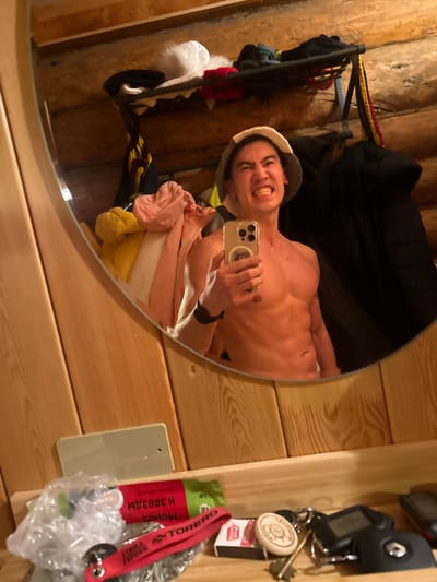 A shirtless man wearing a sauna hat grins at his smartphone while taking a selfie in a round mirror. His toned torso is visible. The mirror reflects a clothing rack with various items. The background shows a wooden wall and a storage organizer containing keys, a lighter, and a "Moroz I Zvezdy" marked item. The entire scene appears to be indoors.