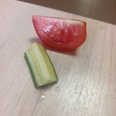 A close-up, high-angle shot captures two sliced vegetables, a tomato wedge and a cucumber piece, resting on a light-colored, wood-grain textured surface. The tomato slice, positioned at the top right of the frame, is a vibrant red with visible seeds and juice glistening on its surface. The cucumber slice, located below and to the left of the tomato, exhibits a pale green rind and a creamy white interior, studded with small seeds. Both vegetables appear fresh, and a small droplet of water is visible on the surface between them. The background consists of the same textured surface, with a blurred area of a darker shade of brown at the top. The scene is well-lit, highlighting the textures and colors of the vegetables and the surface. No people or discernible actions are present. The location is not identifiable from the image.