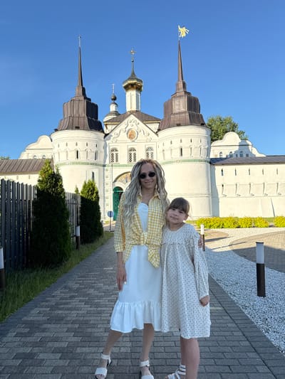 An adult woman and a young girl are standing on a paved walkway, posing in front of the Vysotsky Monastery in Serpukhov, Russia, located near the city of Dumino. The woman, on the left, has long, light blonde hair styled in dreadlocks or similar braids, wears round sunglasses, a white dress with a ruffled hem, a yellow and white gingham plaid shirt tied at her waist, and white sandals. She smiles towards the viewer, with her right arm around the young girl's left shoulder. The girl, positioned to the right, has brown, shoulder-length hair with bangs and wears a long-sleeved white dress with a small dark polka dot pattern and light-colored sandals. She also looks towards the viewer with a slight smile.
The background features the distinctive white architecture of the Vysotsky Monastery, characterized by multiple towers and domes under a clear blue sky. A central building has a golden onion dome topped with a cross, flanked by two taller, conical spires with dark roofing. The spire on the right is topped with a golden angel figure. The white walls of the monastery are adorned with decorative elements, including arched windows, green accents, and a central arched entrance with a green door and what appears to be a religious fresco or mosaic above it.
The paved walkway in the foreground is made of rectangular grey pavers. To the left of the walkway, there is a dark metal fence and several neatly trimmed, conical evergreen bushes. To the right, the walkway borders an area covered in white gravel and lined with low, bright green-yellow bushes, with a dark pole-style light fixture visible. The scene is brightly lit by natural sunlight, suggesting a clear day.