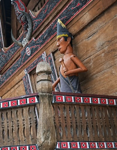 The image captures a close-up view of a traditional wooden building in Ambarita, Indonesia, likely part of a Batak cultural complex. The building features intricate geometric patterns in red, black, and white along its eaves and decorative panels. Prominently displayed on the upper left of the frame are a pair of large, curved water buffalo horns mounted on the wall, a common decorative and symbolic element in Batak architecture.
In the foreground and to the right, a carved wooden effigy of a person stands on a balcony or raised platform. The effigy is depicted with a conical hat adorned with yellow fringe, and it holds a slender object in its hands, resembling a staff or spear. The wooden figure has articulated arms and a visible neck joint. The balcony itself is constructed of carved wooden railings with a repeating decorative motif of red and white squares containing black diamond shapes, set against a wooden background with carved, elongated openings. A large, weathered wooden post is visible on the left side of the balcony, adding to the rustic aesthetic. The overall scene is one of cultural heritage and traditional craftsmanship.