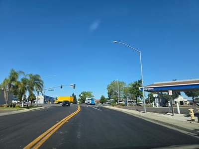 This media file captures a bright, sunny day in West Sacramento, California, likely in the late morning or early afternoon given the strong sunlight and clear blue sky. The scene is a wide, asphalt road with double yellow lines indicating no passing. On the left side of the road, palm trees line the sidewalk in front of a commercial building with an "Amazon" logo visible. Further down the road on the left, a yellow truck is stopped at an intersection, with a traffic light showing green.
The right side of the road features a Chevron gas station with its distinctive blue and white canopy. Several gas pumps are visible under the canopy, and a "NO PARKING" sign is posted. Adjacent to the gas station, other commercial buildings are visible, including one with a "Taco Bell" logo. A bright yellow fire hydrant stands on the sidewalk.
In the mid-ground, a blue tow truck is parked, and other vehicles are scattered in parking lots. The overall impression is a typical suburban commercial area on a clear, pleasant day. There is no indication of people actively engaged in any particular activity, but the presence of vehicles and businesses suggests normal daytime operations. The clear weather and abundant sunlight create a cheerful and ordinary atmosphere. Visible text includes "Chevron," "Amazon," "SMOG," "Harbor Blvd," and "Taco Bell."