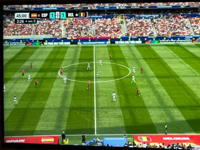 The image shows a live broadcast of a soccer match, likely a FIFA World Cup game given the branding visible on screen. The score is displayed as 1-1 between Spain (ESP) and Belgium (BEL). The game clock shows 45:00 with an additional 2:26 of stoppage time. Players in red and white kits are visible on a green, striped soccer field. The stadium seating is filled with spectators, predominantly wearing red and white attire. The overall environment suggests a professional sporting event. The scene does not provide any information about the city of Vysokaya Gora, Russia, or any specific location within it.