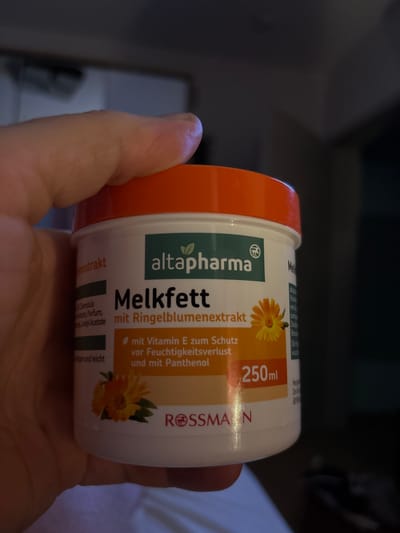 A hand is holding a plastic jar of "Melkfett mit Ringelblumenextrakt" (milk fat with marigold extract) from the brand altapharma. The jar has an orange lid and a white body with text and images printed on it. The text indicates that the product contains Vitamin E for moisture protection and Panthenol. There is an image of a marigold flower on the label. The volume of the product is 250ml. The Rossmann logo is also visible on the jar. The background is dark and out of focus, suggesting an indoor setting. There is no discernible information about the location in Spelle, Germany.