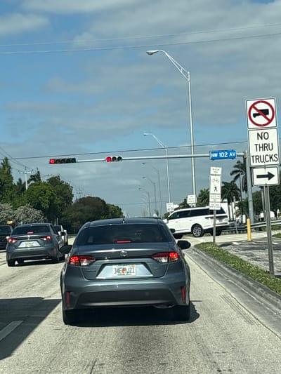 The image captures a daytime street scene in Doral, United States of America, likely from the perspective of a vehicle stopped in traffic. In the foreground, a dark gray sedan (license plate "34E JZI") is visible, with its rear lights and bumper clearly in view. Another similar vehicle follows closely behind. All vehicles appear to be stopped at a red traffic light, which is prominently displayed overhead with three red circular lights illuminated.

The sky is bright but overcast. Palm trees and other green foliage line the side of the road, which features a concrete barrier separating it from a grassy median. Various street signs are mounted on poles above and to the right of the road. Visible text on these signs includes "NW 102 AV" (a street name), "NO THRU TRUCKS" with an arrow pointing straight ahead, and a corresponding circular sign with a red slash over a truck symbol. Further down, signs indicate "BEACON TRAIL," depicting a bicycle and a pedestrian, suggesting a multi-use path. Another sign, partially visible, reads "SPEED LIMIT 40." The license plates on the cars are Florida plates. Shadows cast by the cars indicate the presence of sunlight despite the cloud cover.