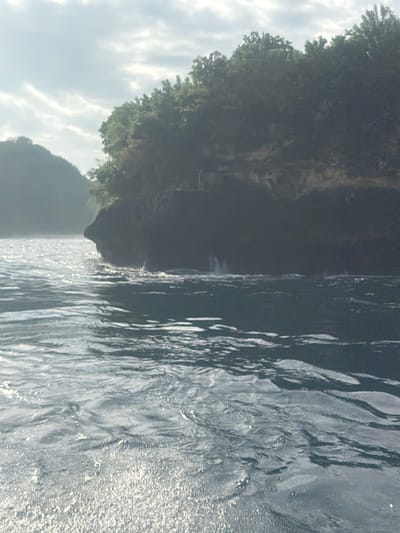 The scene, located in Sakti, Indonesia, features a body of water in the foreground and midground with an agitated surface showing distinct ripples and prominent glints of reflected light. A large, dark, rocky landmass, likely a cliff or island, occupies the right side of the frame, rising steeply from the water. This landmass is densely covered with green tropical vegetation on its upper sections. Multiple narrow streams of water are observed cascading down the cliff face, impacting the larger body of water below and creating visible splashes. A small, indistinct light-colored structure is discernible on the upper portion of this landmass, positioned near the tree line. A second, hazier landmass is present in the distant background to the left. The sky above displays scattered white and grey clouds. No individuals or vessels are present in the image.
