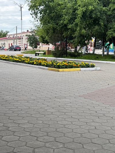 The image shows a street scene in Qarabalıq, Kazakhstan. In the foreground, a paved area with hexagonal gray tiles stretches out. To the left, a flower bed filled with yellow and orange marigolds borders the paving. Behind the flower bed, a low stone wall separates the flowers from a grassy area. Beyond the grass, a wooden bench with metal supports is visible. A black car is parked on the road to the left. In the background, a long, light-colored building with red trim and air conditioning units is visible. Several trees with green foliage are scattered throughout the scene, some with whitewashed trunks. A utility pole with wires stands near the building. The sky is overcast.