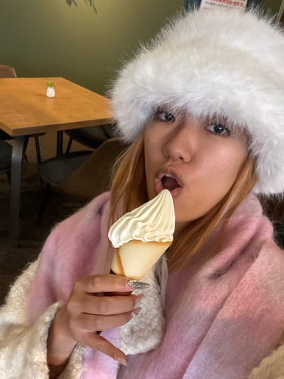 A young woman wearing a fluffy white hat and pink sweater holds a pointed dessert with white frosting close to her open mouth. Her long, ornate nails are adorned with gemstones. She appears to be indoors, seated near a wooden table with a small floral decoration. Her light brown hair is visible beneath the hat. Her expression is one of excitement or anticipation.