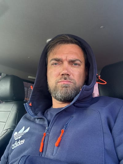 A middle-aged Caucasian man with a beard is taking a selfie inside a vehicle. He is wearing a dark blue hooded Adidas jacket with orange zipper pulls, and a dark blue t-shirt underneath. The interior of the vehicle is visible, including dark leather seats. The man's expression is neutral, and his eyes are looking directly at the camera. The lighting suggests it is daytime. The image appears to be taken in Usinsk, Russia, based on the metadata.