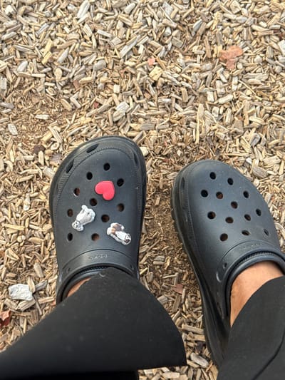 A bird's-eye view shows black Crocs on wood chips. One Croc is adorned with a red heart and two dog-shaped charms. Black leggings are visible around the ankles. The wood chips vary in color from light tan to brown and cover the ground.