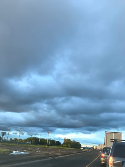 The media file shows a view of a road with traffic, under a dramatic sky. The scene appears to be in Валы, Russia. A line of vehicles, including cars and semi-trucks, is stopped on a two-lane road. The sky is filled with dark, ominous storm clouds, interspersed with patches of brighter, lighter blue sky, suggesting an impending storm or the aftermath of one. The time of day is likely late afternoon or early evening, given the lighting and cloud formations.
The foreground shows a dark asphalt road with a yellow dividing line. To the left of the road, there is a grassy area with some puddles of water, reflecting the sky. Several utility poles with wires are visible along the side of the road, and in the distance, a line of trees forms the horizon. On the right side of the road, the back of a dark SUV is visible, with its taillights illuminated. Further down the road, a large white semi-truck with "KOLLMAN" written on its trailer is prominent.
The overall mood conveyed is one of anticipation or a slight sense of unease due to the stormy weather, juxtaposed with the mundane reality of a traffic jam. There is no visible text other than the brand name on the truck.