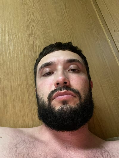 A man with a full beard and dark hair is shown from the chest up, looking directly at the camera. He has brown eyes and fair skin with some redness on his cheeks. The background is a light brown wooden panel wall. The image appears to be a selfie taken indoors. There is no visible indication of the location within the city of Unknown, Bangladesh.