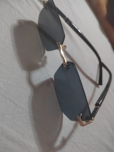 A pair of dark-lensed, frameless sunglasses with gold accents lies on a white, textured surface, casting a shadow. The sunglasses are positioned diagonally, with the left lens closer to the camera and slightly angled upwards. The right temple arm extends upwards and to the right, with a subtle bend. The surface appears to be fabric, possibly a tablecloth or bedsheet, with a visible weave pattern. The lighting suggests an indoor setting with a light source casting a distinct shadow of the sunglasses onto the surface. There are no people or other objects visible in the image. The location is not specified in the image itself, and therefore it is not possible to determine if it is in Dorkovo, Bulgaria.