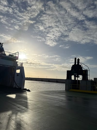 The image depicts a scene on a vessel in Yaiza, Spain, potentially a ferry or a large boat, at dawn or dusk given the low sun and cloudy sky. To the right of the frame is a section of the vessel's deck, appearing wet or polished. A piece of heavy machinery is visible, possibly part of the vessel's equipment. A coastline is discernible in the distance with what appears to be buildings or structures lined along the coast. To the left are white cumulus clouds. In the foreground are sections of the vessel, including railings and life-saving equipment.