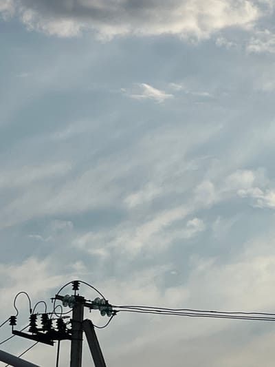 An outdoor, medium shot captures a utility pole with electrical equipment against a cloudy sky in Golubitskaya, Russia. The pole, made of concrete, supports multiple insulators and power lines. The sky is filled with scattered clouds, some appearing wispy and others denser, with a pale blue hue visible in between. The scene is devoid of any people or animals. The focus is on the infrastructure against the natural backdrop of the sky.