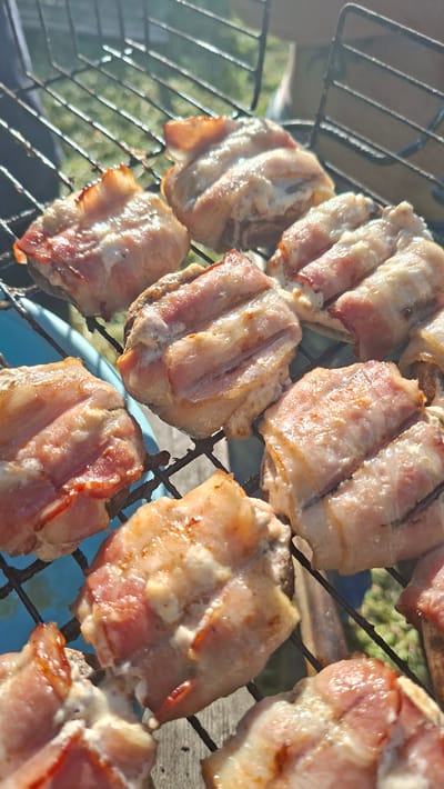 The image depicts several pieces of bacon cooking on a metal grill. The bacon is arranged in a grid pattern and appears to be in the process of being cooked. The environment visible behind the grill suggests an outdoor setting, possibly a backyard or picnic area. The location is Сваткаўскі сельскі Савет, Belarus. No people are visible in the image.