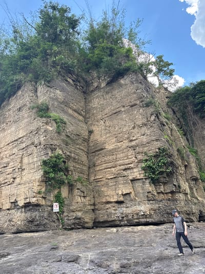 The image depicts a rugged cliff face with a person walking on rocky terrain in the foreground. The cliff is composed of layered rock, with vegetation, including trees and vines, growing from its crevices and from the top. A red and white sign, with Vietnamese text, is visible on the cliff face. The sky is blue with some white clouds. The person, wearing a gray t-shirt, black pants, a cap, and athletic shoes, is walking away from the camera towards the right side of the frame. The environment suggests a natural, outdoor location, possibly a hiking or scenic area within Tân Phú, Vietnam.