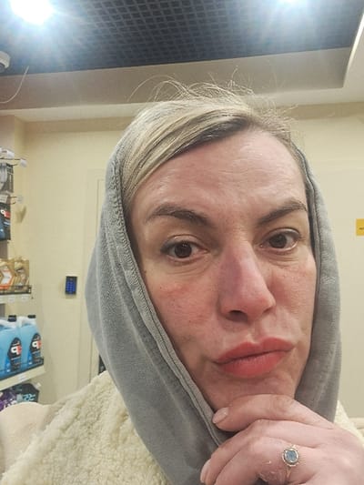 A fair-skinned woman with light blonde hair and a gray hoodie is captured in a close-up shot. She has brown eyes and red lips. Her right hand is visible, displaying a ring with a blue stone. The background reveals a wall, shelves with products, and a portion of a window or screen reflecting light.