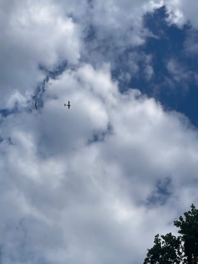 The image captures a view of the sky on a cloudy day, with two small airplanes flying. The sky is a mix of blue and white, with large, fluffy cumulus clouds. The planes are silhouetted against the clouds, appearing as small dark shapes. The top right corner of the image shows the dark green leaves of a tree, suggesting an outdoor setting. The lighting indicates it is daytime, likely afternoon given the cloud formations. The scene is peaceful and serene, with the planes appearing to be engaged in recreational flying. There is no visible text in the image. The location context of Schrecksbach, Germany, suggests this scene is taking place in a rural or semi-rural area, which aligns with the presence of open sky and trees.
