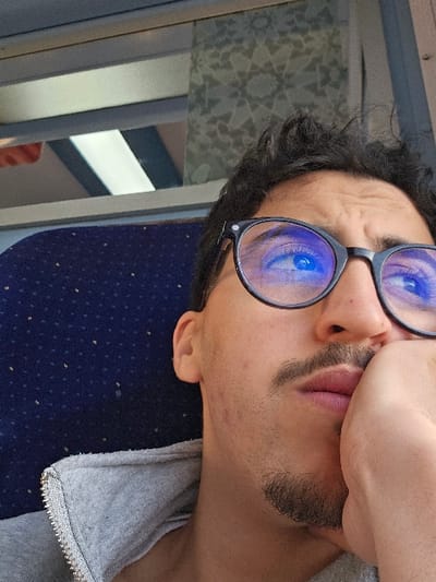 The scene is situated in the city of Ouahat Sidi Brahim, Morocco, depicting a male individual in a close-up, slightly upward-angled shot. The subject has dark, curly hair, a mustache, and a short beard. He wears black-framed glasses, where a distinct blue reflection is visible across the lenses, obscuring the eyes. His gaze is directed upwards and to the right. The individual's left hand is positioned against his lower chin and mouth area, with fingers curled, indicating a contemplative or resting posture. He is attired in a light grey hooded sweatshirt with a visible silver-colored zipper on the collar. The background features a dark blue upholstered seat, patterned with numerous small, light-colored dots, positioned to the left of the subject. Above the individual's head, a window structure with a grey frame is partially visible, alongside a light-colored panel displaying a repeating geometric design.