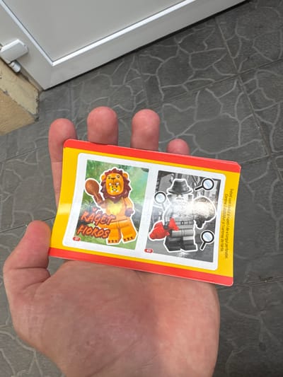 A hand holds a rectangular card with a red border and a yellow background. The card is divided into two sections. The top section features a black and white illustration of what appears to be a toy set, with magnifying glasses highlighting different elements. There are also some red and grey toy pieces visible. The bottom section of the card displays a colorful illustration of a Lego-style lion character. The lion is orange with a brown mane and is set against a green, leafy background. Text is visible on the card, including "RAGET SOYOH" above the lion illustration and a small number "37" to its right. In the top yellow area, there is text in Romanian that translates to "Fold the card easily and tear it along the perforation. Contains 2 stickers and 1 paper card." A small "43" is visible to the right of the top illustration. The background shows a tiled surface with a distinctive pattern. The scene is outdoors, with a white framed object, possibly a window or door frame, visible on the left side. The lighting suggests it is daytime. The hand holding the card appears to be an adult's, with visible fingerprints on the card and the thumb positioned to hold the bottom edge. The overall impression is of a game or collectible card.