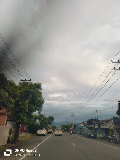 The image depicts a street scene in Teungoh Baro, Indonesia on May 23, 2026, at 18:21 local time. The sky is overcast with heavy clouds. A paved road stretches into the distance, with a white car and a silver car moving along the road. A motorcycle is further ahead. Buildings and trees line the roadside on both sides. Electrical power lines run overhead. Several parked motorcycles are visible on the side of the road.