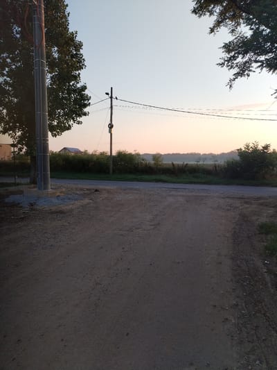 The image depicts an outdoor scene, likely at dawn or dusk, in Moara Domnească, Romania. A gravel road occupies the foreground, leading towards a paved road in the midground. On the left side of the paved road stands a tall, grey utility pole with visible wires attached. To the right of this pole, a second, shorter utility pole is visible, also with wires and a circular object suspended from it. A bird is perched on one of the wires near the second pole. Beyond the paved road, a line of dense green foliage stretches across the landscape, with trees and bushes. In the background, a house with a red roof is partially visible through the trees, and further in the distance, a hazy, treed horizon is apparent under a pale, lightly colored sky. The overall environment appears rural or semi-rural.