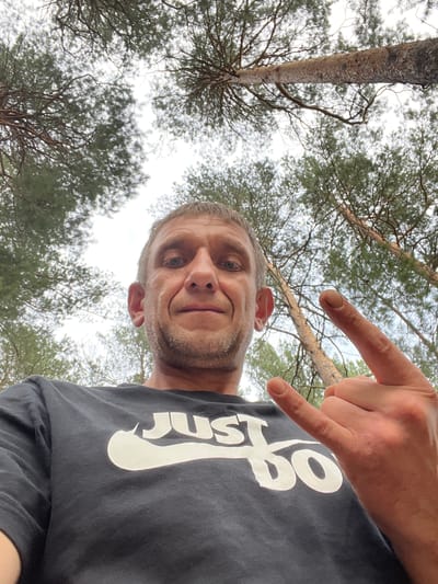 A man in a black t-shirt with the "Just Do It" logo is making a rock-and-roll hand gesture while looking up at the camera from a low angle. He is in a wooded area with tall pine trees overhead and a bright, overcast sky. The man has short, graying hair and stubble on his face. His expression is neutral. The image appears to be a selfie taken outdoors. There is no indication of the city of Yuzhnyy, Russia, in the image.