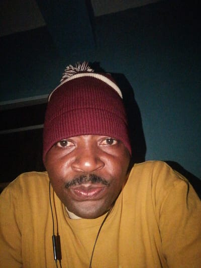 A man wearing a maroon beanie with a white and maroon pom-pom, and a mustard yellow t-shirt, is captured in a close-up, front-facing shot. He has a mustache and appears to be wearing earbuds. The background is a dark, teal-colored wall. The lighting is dim, with shadows casting across the scene. The image was taken in Ajasse Ipo, Nigeria.