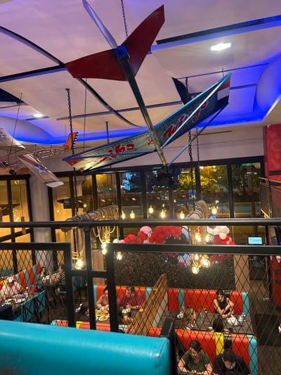 The image depicts an interior setting. A decorative airplane model hangs from the ceiling. Below, a multi-level dining area features people seated at tables. String lights and red and white balloons adorn the space. A metal grid partially obscures the view of the diners, creating a layered perspective. The scene appears to be a restaurant or entertainment venue.