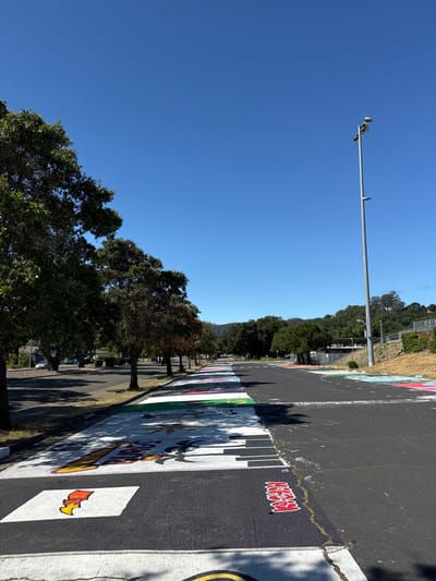 The image captures a parking lot in Novato, California, USA, on a clear, sunny day. The pavement is decorated with colorful artwork, resembling a piano keyboard and various other designs, painted across the parking spaces. Trees line the left side of the parking lot, with shadows cast on the ground. In the background, a tall light pole stands, and hills covered in trees are visible. The scene appears to be an outdoor public space, possibly a school or community center parking lot, given the vibrant murals.