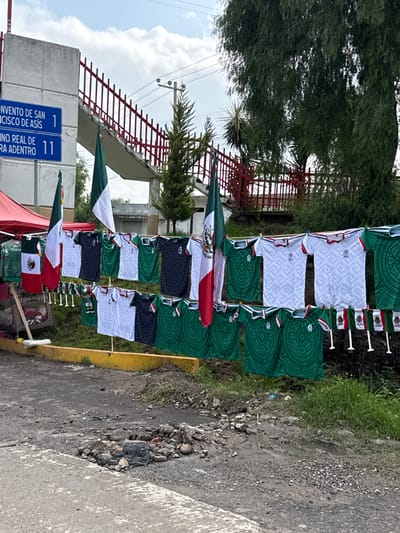 The image depicts a street scene in Tepeji de Ocampo, Mexico, where merchandise, specifically shirts and small flags, are displayed for sale. The items prominently feature the colors and symbols of the Mexican flag and indigenous patterns, suggesting a patriotic or cultural theme. These goods are hung on a clothesline, with a red tent visible in the background on the left, likely part of a vendor's setup.

A concrete structure with blue street signs is present on the left side of the frame. One sign indicates "CONVENTO DE SAN FRANCISCO DE ASIS" with the number "1," and another reads "CAMINO REAL DE LA SIERRA ADENTRO" with the number "11." These signs provide specific geographical context within Tepeji de Ocampo.

In the background, a red metal railing lines a raised walkway or staircase, with lush green trees and vegetation surrounding the area. The ground in the foreground is a rough, unpaved surface with scattered rocks and debris, including a pile of stones and what appears to be a black trash bag. The sky above is partly cloudy. No people are clearly visible in the image, but the setup implies the presence of vendors and potential customers. The overall impression is of an outdoor market or roadside stall selling nationalistic or culturally themed items in a semi-urban or suburban setting.