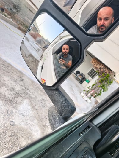 A man with a shaved head and a beard, wearing a grey t-shirt, is reflected in a large side-view mirror of a white truck. He is holding a smartphone in his left hand and appears to be taking a selfie. The truck is parked on a gravel surface, possibly in an industrial or construction area, with piles of reddish-brown material and some building structures visible in the background. In the foreground, part of the truck's interior, including the dashboard and door panel, is visible. The overall scene suggests a sunny day. The image is cropped in such a way that the truck's mirror and a portion of the vehicle are prominently featured. The man's reflection is also visible in a smaller inset mirror. The location is identified as Nàquera / Náquera, Spain, implying a likely Spanish setting.
