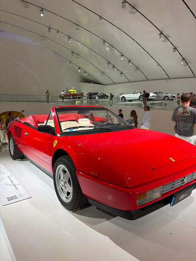 The image shows a red Ferrari Mondial Cabriolet on display in a museum. The car is positioned in the foreground, angled slightly towards the left of the frame. The interior of the car is visible, with beige seats and red upholstery. A plaque with the car's details and a QR code is visible on the floor to the left of the car. In the background, other cars are on display, including what appears to be a white convertible and a light gray convertible, as well as a brown car with a yellow roof. Several people are present in the background, observing the cars. The environment appears to be a modern, spacious museum with a white, curved ceiling and track lighting. The floor is a polished white surface. The plaque in front of the Ferrari reads "FERRARI MONDIAL CABRIOLET". The museum's name is visible on a sign attached to the front of the Ferrari: "MUSEO ENZO FERRARI MODENA". This indicates the location is the Enzo Ferrari Museum in Modena, Italy.
