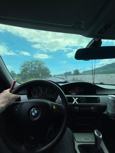 A first-person perspective shot from inside a BMW car. The dashboard, steering wheel with the BMW logo, and instrument panel are visible. The driver is holding a pen-like object. Outside the car, the view shows a blue sky with clouds and green trees. A road with a fence running along it can be seen. The car's center console has climate control buttons. An energy drink is to the side of the console, and the gear shift is visible. The location might be near a road with fencing and vegetation in Belovo, Bulgaria, based on the landscape visible through the windshield.