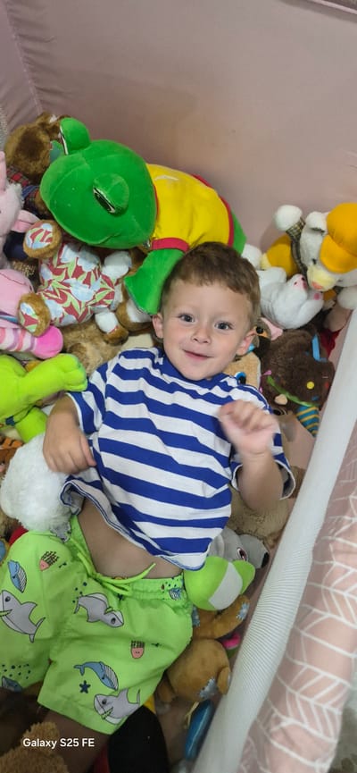 A young boy with brown hair and blue eyes lies on his back amidst a collection of stuffed animals. He is wearing a blue and white striped t-shirt and bright green shorts with a shark print. The boy is smiling and looking up at the camera. The stuffed animals include a large green frog, a yellow and red turtle, and various teddy bears and other plush toys. The scene appears to be indoors, possibly in a playpen or bed. The background shows a pink fabric with a leaf pattern. There is no indication of the location being in Almáchar, Spain.