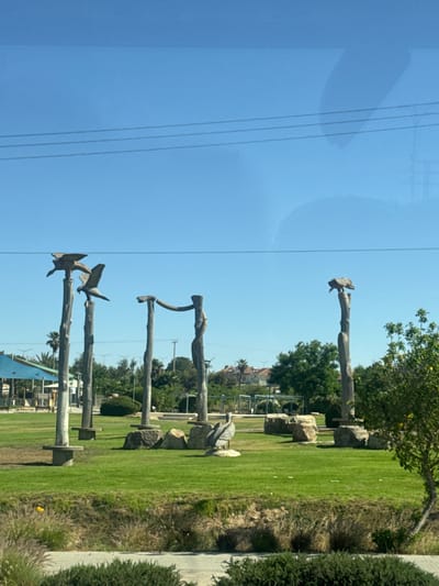 The image shows a row of wooden sculptures in a park setting. These sculptures appear to be abstract representations of animals or natural forms. They are placed along a grassy area adjacent to a paved walkway and some shrubbery. In the background, there are trees and what appears to be the outskirts of a residential or commercial area. The sky is clear and blue. The location is in Arad, Israel, as the sculpture park "Omanut BaRuach" (Art in the Wind) features similar wooden sculptures and is located in that city.