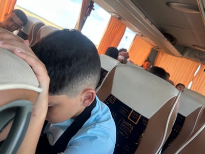 The image shows the interior of a bus, with several people visible. The subjects are primarily young males, seated on the bus. One person in the foreground is wearing a light blue t-shirt and has short dark hair. Another person is seen in the background, also with dark hair. The bus seats are brown and white, with a patterned fabric visible on the seat backs. The windows of the bus are covered with orange curtains. The lighting suggests it is daytime, possibly sunny, as light filters through the curtains. The people appear to be resting or looking out, with one individual's head resting on their arm. The overall scene depicts a journey or travel situation. There is no visible text in the image.