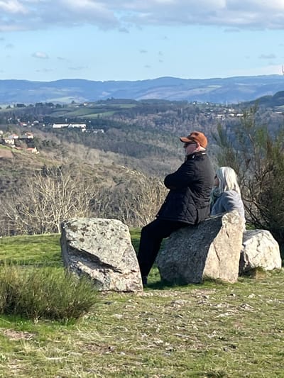 A daytime image depicts two individuals seated on large, weathered grey rocks atop a grassy hill overlooking a valley in Chambles, France. The male figure on the left is positioned with his back partially to the camera, wearing a brown baseball cap, dark sunglasses, a black quilted jacket, and dark trousers, with his arms crossed. To his right, a female with long blonde hair, dressed in a light grey top, is seated on an adjacent rock, also facing the distant landscape. The foreground consists of patchy green and brown grass with sparse low-lying vegetation. The background features a vast natural landscape under a partly cloudy blue sky, encompassing a valley with sparse deciduous trees, scattered residential structures, and several layers of distant blue-hued rolling hills extending to the horizon.