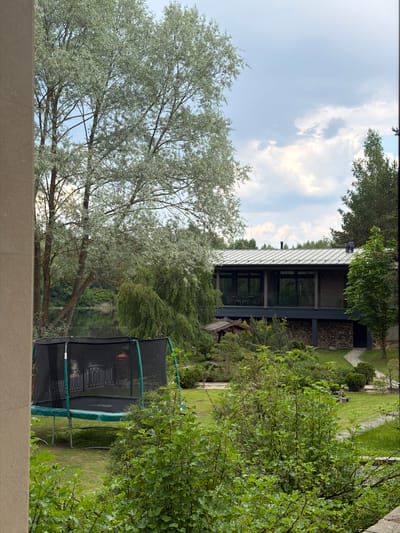 The image shows a residential scene in Shchelkovo, Russia. In the foreground is a large tree with silvery foliage. A green and black trampoline is visible on the right side of the image, situated on a lawn. In the background, a modern house with a gray roof and large windows is partially obscured by trees. The sky is visible with some clouds. The scene includes elements of a well-maintained yard and the architecture of a contemporary home within a lush, green environment, typical of suburban or semi-rural areas.