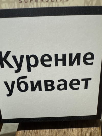 The image shows a close-up of a white surface with large black Russian text. The text reads "Курение убивает" which translates to "Smoking kills". The letters are bold and sans-serif. The surface appears to be part of a cigarette pack, as indicated by the text and the black border around the white area. There is also a hint of text in a lighter color above the main text, which appears to say "SUPER SLIMS". The background is out of focus but suggests a wooden surface beneath the pack. The image does not contain any information about the city of Tsandrypsh, Abkhazia.
