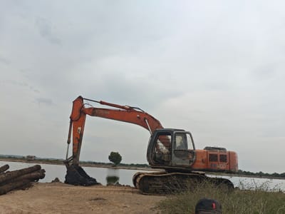 In the city of Kualaperak, Indonesia, a weathered, orange Hitachi EX 200 excavator rests on a dusty embankment beside a calm body of water. The excavator's boom is extended, and its bucket is filled with dark, wet soil. To the left of the excavator, a pile of rough-hewn logs lies on the ground. In the distance, across the water, a small, rustic wooden structure with a red roof stands near the treeline, and a solitary tree is visible in the middle of a field. The sky is overcast, casting a diffused light over the scene. The foreground is dominated by dry earth and sparse vegetation. A person's black cap is visible in the lower right corner of the frame, suggesting a human presence at the site.