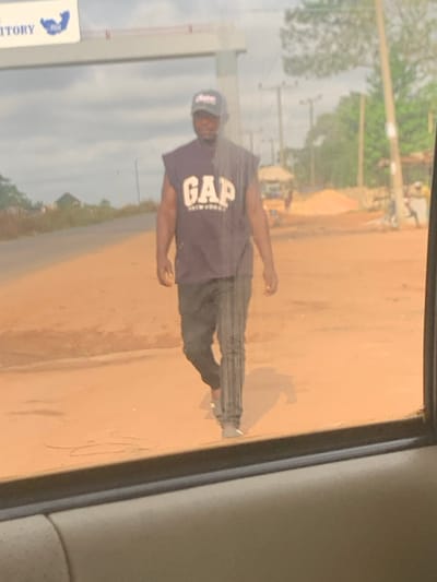 The image depicts an outdoor scene in Issele Uku, Nigeria, captured from inside a vehicle through a window, evidenced by the vehicle's window frame at the bottom and vertical streaks on the glass.
The central subject is an adult male, of apparent Nigerian ethnicity, walking towards the viewer on an unpaved, reddish-brown dirt path alongside a paved road. He is dressed in a dark baseball cap, a dark sleeveless shirt with "GAP NEW YORK" emblazoned in white across the chest, dark trousers (possibly track pants), and light-colored sneakers. His right arm is slightly bent, and his left arm hangs straight down.
In the background, on the left side, an overhead structure is visible, featuring partial text "TERRITORY" and "NIGERIA" with a blue map graphic. To the right, utility poles, scattered trees, and a pile of reddish-brown sand or earth are present. Further in the distance, a light-colored structure and a yellow tricycle (keke napep) are discernible. The sky is overcast with light grey clouds. The overall environment suggests a rural or semi-urban roadside setting.