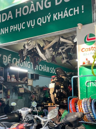 This is a medium shot of a motorcycle repair shop in Liên Nghĩa, Vietnam. The shop is cluttered with motorcycle parts hanging from the ceiling and shelves stacked with various items. A green sign above the entrance reads "IDA HOÀNG Đ” and "ANH PHỤC VỤ QUÝ KHÁCH!". Below this sign, another green sign says "ĐẾ CHÚNG TÔI CHĂM SÓC". To the right, a white sign with a green and red logo displays "Castrol Bike Po" and "Cha".

In the foreground, two motorcycles are visible, one white and one red. A person in a green shirt is working on a motorcycle in the middle of the frame. Several tires are neatly stacked in a rack to the right. The walls are a light blue and adorned with various tools, a clock, and shelves filled with more items. The overall impression is of a busy, working motorcycle repair shop.