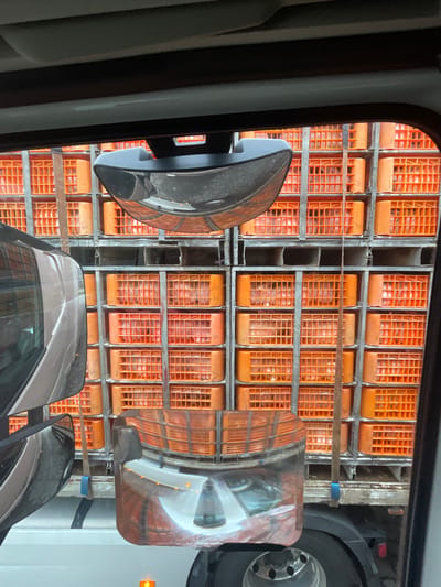 A forensic view from within a vehicle in Chaves, Portugal, depicts the rear of a commercial flatbed truck loaded with numerous bright orange, perforated plastic crates. These crates are stacked vertically and horizontally in multiple tiers on gray metal racks and pallets, completely filling the visible cargo area. Each crate holds numerous pale, ovoid produce items, likely onions, visible through the perforations. An upper convex mirror and a lower reflective surface project distorted views of the roadway and surrounding reflections. A partial reflection of the originating vehicle's silver-toned side panel and window is evident on the left. A wooden pole is vertically secured to the right side of the truck's load structure by a blue strap. A segment of a truck tire and a small amber marker light are visible at the bottom of the frame.