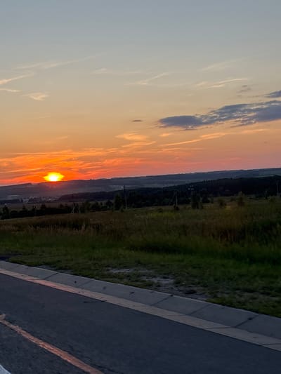The image shows a sunset over a rural landscape, likely in or near Mayevka, Russia. The sun is a bright, intense orange, low on the horizon, casting a warm glow across the sky which transitions from orange to pale blue. Wispy clouds are scattered across the sky. The foreground features a dark asphalt road with a faded orange line, bordered by a concrete curb and grass. Beyond the road, a field of dry, golden grass stretches towards a distant line of dark trees. In the middle ground, hills are visible, dotted with small structures suggesting a village or scattered settlements. The overall impression is one of tranquility and the end of a day in a rural setting.