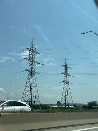 The image shows a daytime view of a roadside scene in Sagholasheni, Georgia. Two tall, lattice-style electricity pylons dominate the frame, with power lines stretching across the sky. A white SUV is parked on the side of the road, partially obscured by a concrete barrier. In the background, several wind turbines are visible on the horizon, suggesting a rural or semi-industrial landscape. The sky is blue with scattered clouds, and the overall lighting indicates it is daytime. The concrete barrier in the foreground suggests the perspective is from another vehicle on a road.